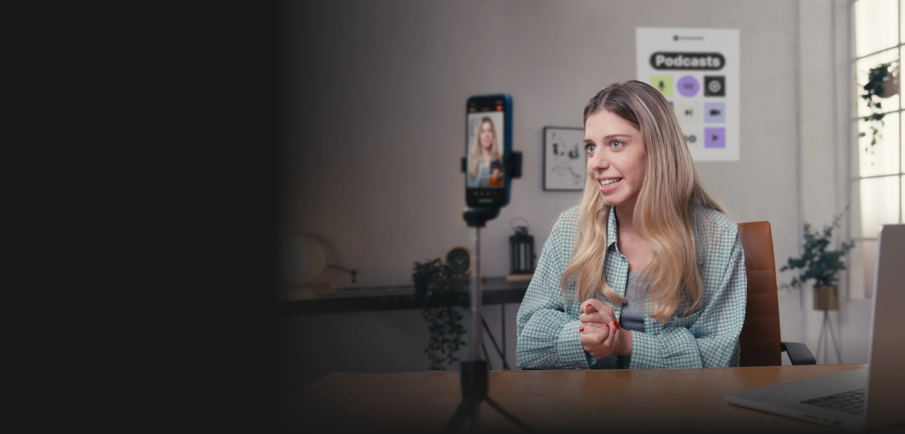 Young woman with long blonde hair speaking during a video call recorded on a smartphone mounted on a tripod, sitting in a bright room with podcast posters on the wall.